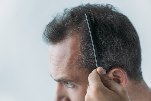 close-up view of mid adult man combing hair with comb isolated on grey, hair loss concept