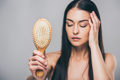 stressed brunette woman with closed eyes holding hairbrush isolated on grey, hair loss concept
