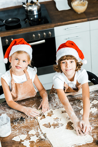 high angle view of adorable kids in santa hats preparing christmas cookies and smiling at camera