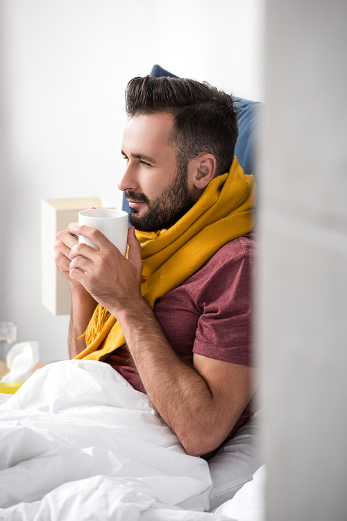 smiling sick young man with scarf holding cup of hot tea while sitting in bed