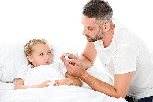 father looking at electronic thermometer while adorable ill daughter with temperature lying in bed, isolated on white