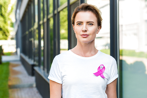 pensive middle aged woman with pink ribbon standing on street, breast cancer awareness concept