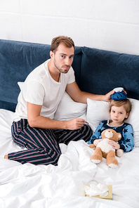 high angle view of father checking temperature of son with electric thermometer and holding ice pack on his head in bed and 