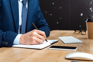 cropped image of businessman writing in textbook at table with smartphone in modern office