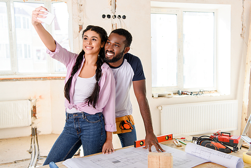 cheerful african american couple taking selfie on smartphone near table with blueprint during renovation of home