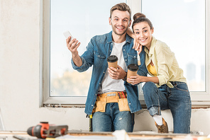 happy young couple with coffee to go and smartphone smiling at camera during renovation