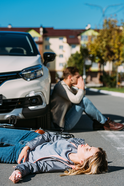 selective focus of injured woman lying on road after car accident with shocked car driver behind