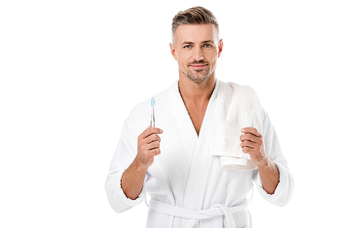 cheerful adult man in bathrobe with towel over shoulder holding toothpaste and toothbrush isolated on white