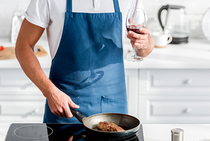 cropped view of man with glass of wine cooking steak on pan
