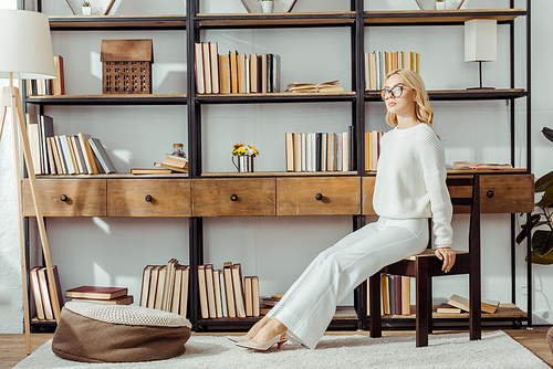 adult blonde woman in glasses sitting near rack with books