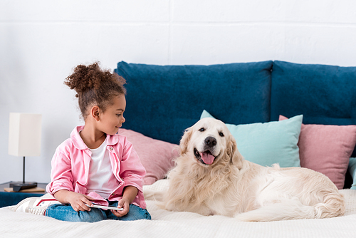 Curly african american kid sitting on the bed with smartphone and smiling golden retriever