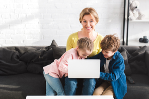 happy mother smiling at camera while using laptop with children at home