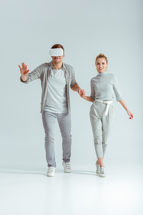 smiling couple holding hands while man experiencing virtual reality on grey background