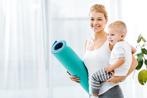 beautiful woman holding blue fitness mat and smiling cute toddler boy