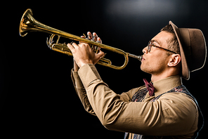 side view of young male jazzman in hat and eyeglasses playing on trumpet isolated on black