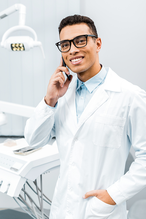 handsome african american doctor in glasses talking on smartphone with hand in pocket