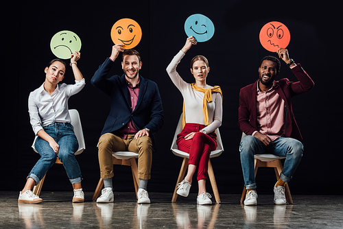 multiethnic people sitting on chairs and holding face cards with various emotions isolated on black