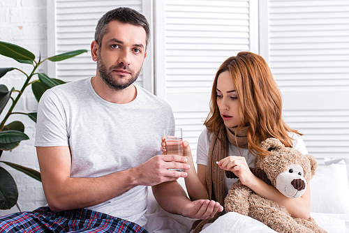 husband giving pills and glass of water to sick wife with teddy bear in bedroom