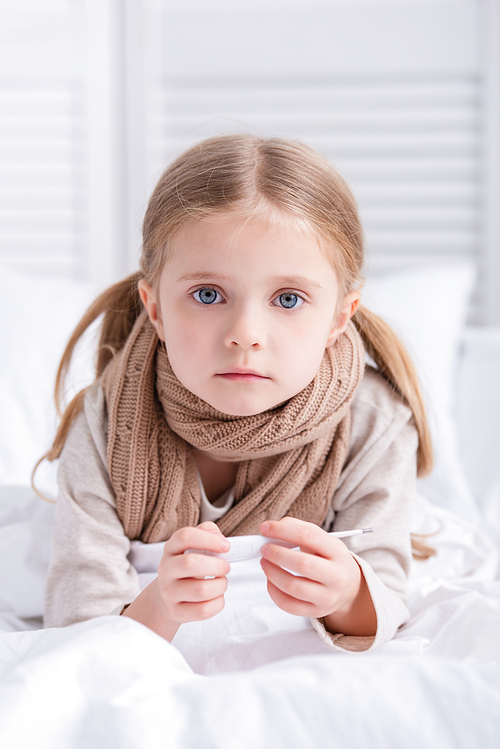 sick child with scarf over neck lying in bed, holding thermometer and  at home