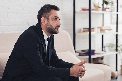 side view of worried handsome businessman in suit sitting on sofa in office and looking away