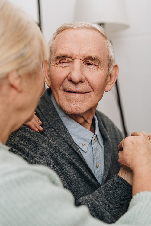 selective focus of smiling pensioner looking at senior wife at home