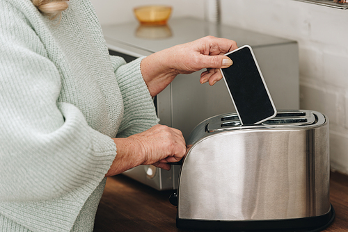 cropped view of retired woman with dementia disease putting smartphone with blank screen in toaster