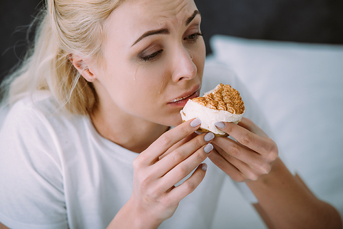 selective focus of depressed woman in pajamas eating cake in bed alone