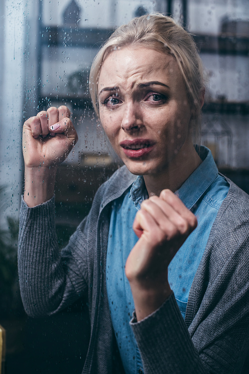 stressed adult woman with clenched fists  at home through window with raindrops