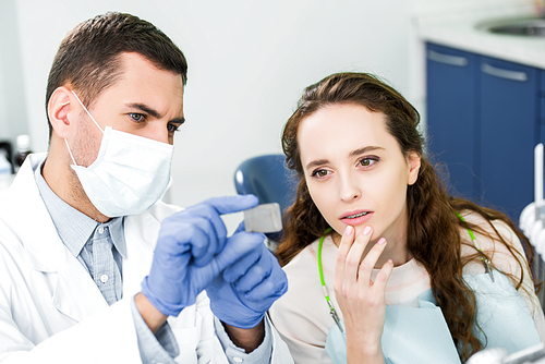 selective focus of pensive woman looking at x-ray of teeth in hands of dentist in latex gloves and mask