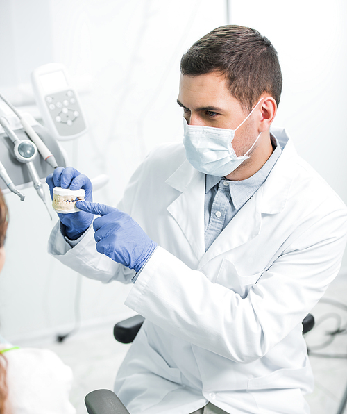 dentist in mask and latex gloves pointing with finger at teeth model near female patient