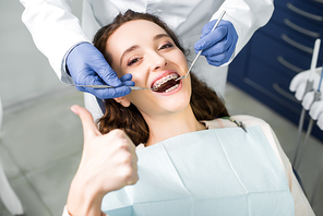 cropped view of dentist in latex gloves examining attractive woman in braces with opened mouth showing thumb up