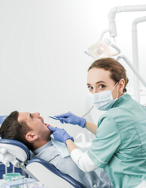 dentist in mask holding dental drill while working with patient in dental clinic