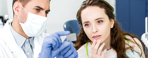 selective focus of thoughtful woman looking at x-ray of teeth in hands of dentist in latex gloves and mask