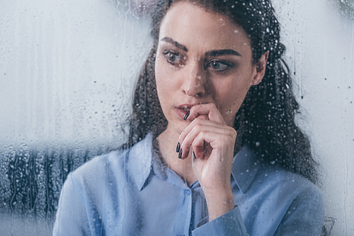 beautiful upset woman touching face and looking through window with raindrops