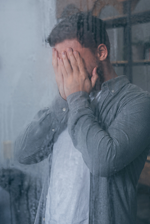 adult man covering face with hands and crying through window with raindrops