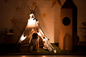 Kid standing on all fours in wigwam and looking at cardboard rocket in dark room