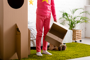 Cropped view of kid holding cardboard helmet and standing near rocket in bedroom