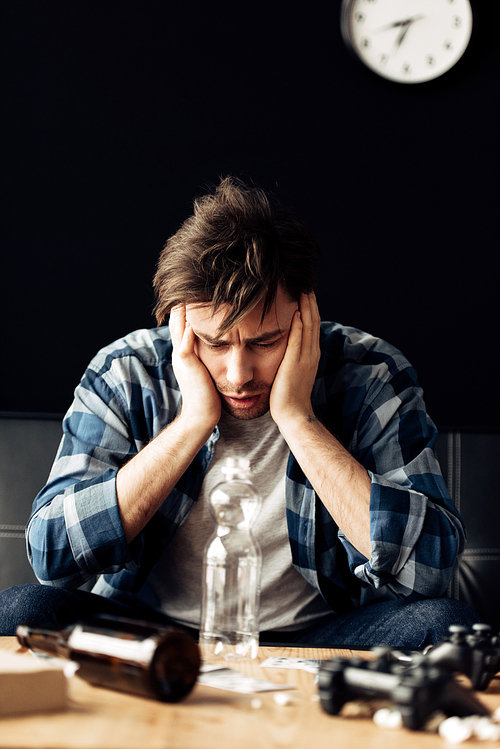 man suffering from hangover holding head and looking at bottle at home