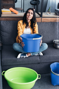 Upset woman with buckets and basin looking at leaking ceiling