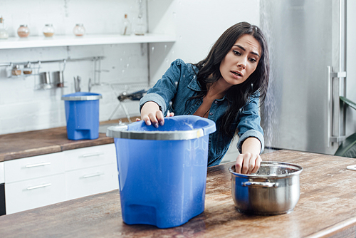 Stressed young woman using buckets and pot during leak in kitchen