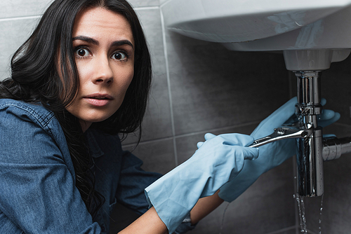 Amazed brunette woman in rubber gloves repairing sink pipe with wrench