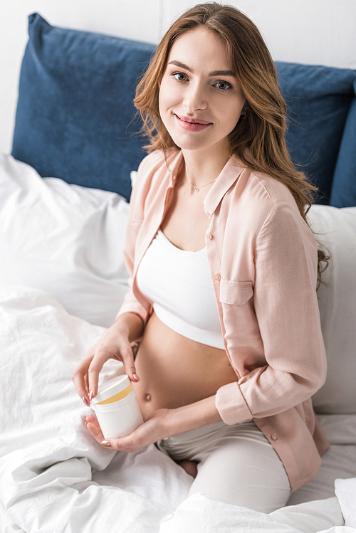 Smiling pregnant woman sitting on bed and opening container with cream