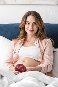 Happy pregnant woman lying in bed with apple and 