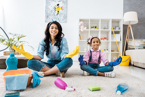 smiling african american mother and daughter in bright rubber gloves sitting on carpet in lotus poses