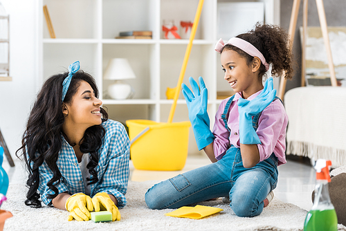 smiling african american mom and daughter in bright rubber gloves talking while resting on carpet