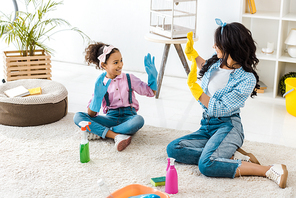 african american mom and cute daughter in bright rubber gloves having fun while sitting on carpet