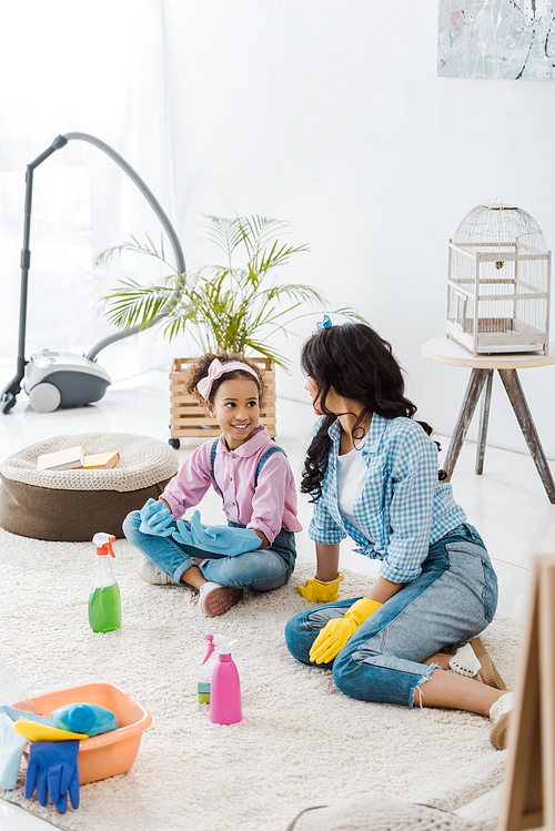african american american mother and daughter in bright rubber gloves talking while sitting on carpet
