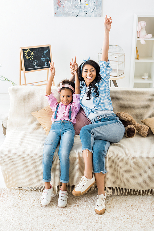 cheerful african american mom and daughter showing victory signs while sitting on sofa at home
