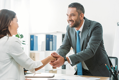 handsome advisor in suit shaking hands with investor at workplace