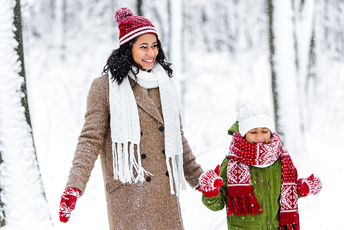 cheerful african american mom and cute child standing during snowfall in winter park
